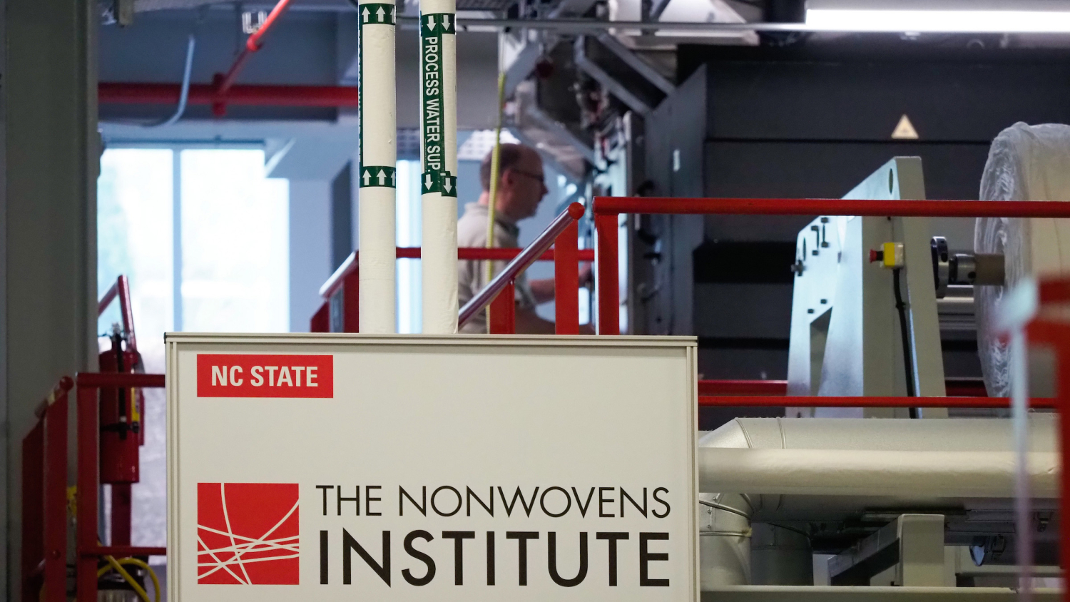 A sign with NC State's logo and "The Nonwovens Institute" on it is visible in front of lab equipment with a person at work.