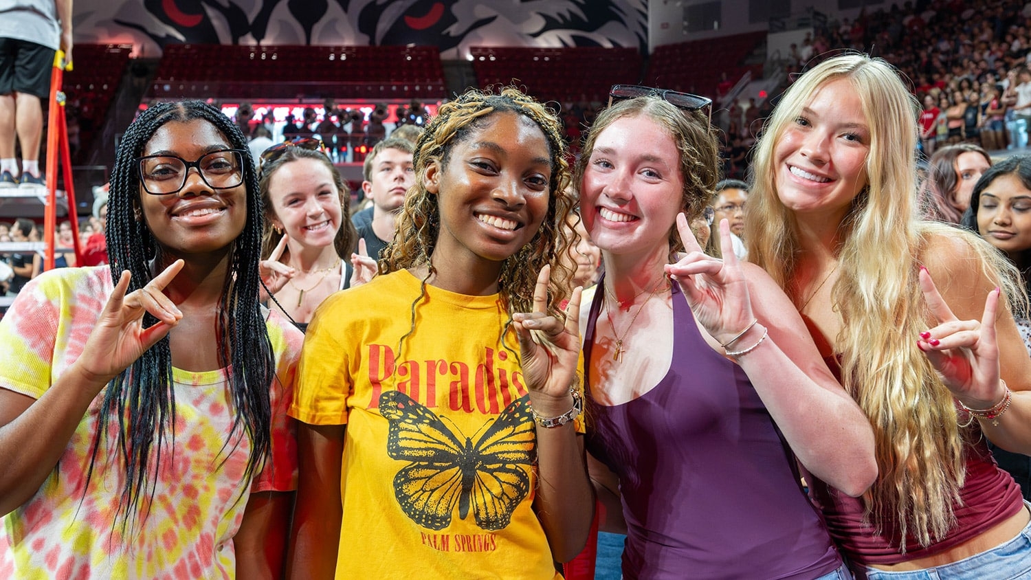 Four students hold up the wolfie hand gesture with a large crowd gathered behind them inside Reynolds Coliseum during new student convocation.